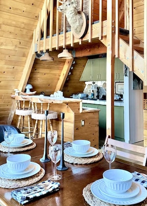 A wooden interior with a dining area set for four, a staircase, and a kitchen visible in the background. - The Highpoint A-Frame | Walkable | BBQ | Max 4 (Big Bear Lake)