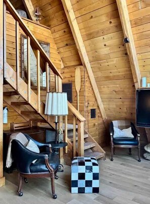 A wooden interior with a staircase, a black leather chair, a checkered ottoman, and a television. - The Highpoint A-Frame | Walkable | BBQ | Max 4 (Big Bear Lake)