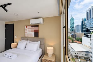 Ein Hotelzimmer mit Bett, Nachttischlampen, einer Klimaanlage, Blick auf die Stadt und einem modernen abstrakten Gemälde.