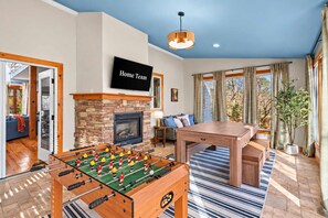 A living room with a foosball table, a fireplace, a television, and a dining area. - Helen Haus: Golf Sim & Hot Tub Theater Retreat (Helen)