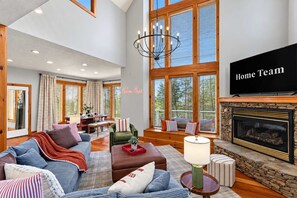 A spacious living room with a fireplace, large windows, and a television. - Helen Haus: Golf Sim & Hot Tub Theater Retreat (Helen)