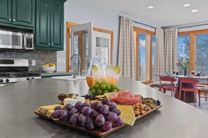 A kitchen island with a fruit and cheese board, a pitcher of iced tea, and a dining area with a table and chairs. - Helen Haus: Golf Sim & Hot Tub Theater Retreat (Helen)