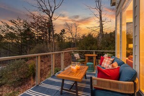A balcony with a wooden table, chairs, and a sofa with cushions. - Helen Haus: Golf Sim & Hot Tub Theater Retreat (Helen)
