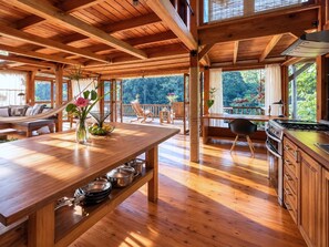 A spacious wooden interior with a dining area, kitchen, and seating by a large window overlooking greenery. - Kubla Khan - Chirripó riverfront mountain retreat (Perez Zeledon)