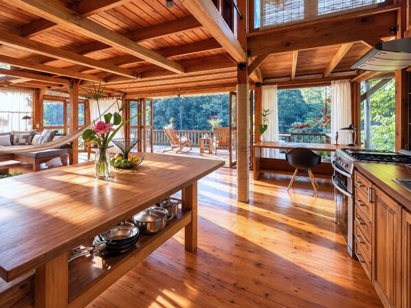 A spacious wooden interior with a dining area, kitchen, and seating by a large window overlooking greenery.