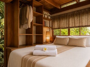 Ein Schlafzimmer mit Bett, Kleiderschrank, einer Bücherablage und einem Fenster mit Blick auf Grünflächen.