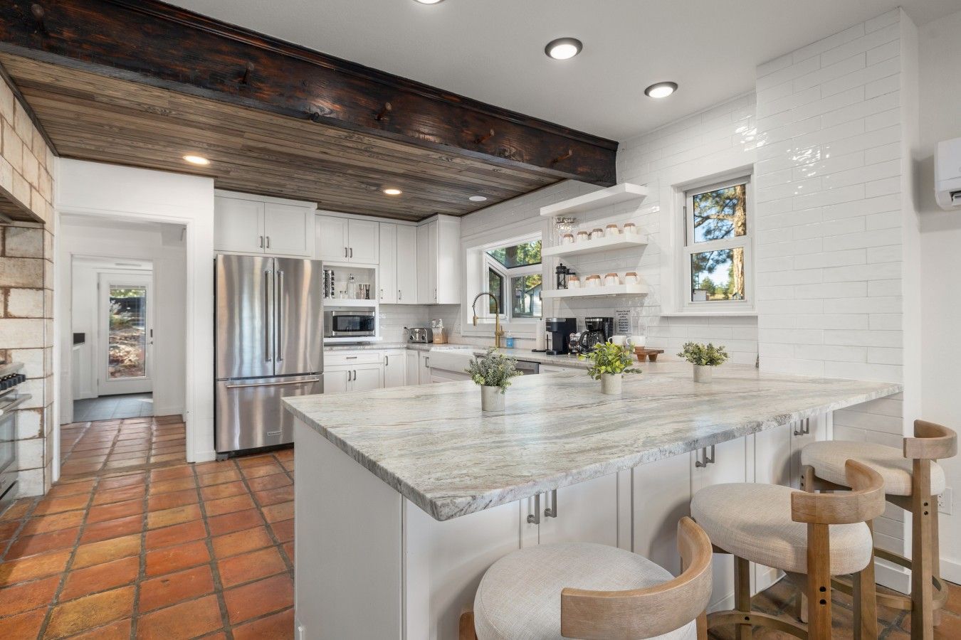A modern kitchen with white cabinetry, a large island with bar stools, a stainless steel refrigerator, and a wooden ceiling.
