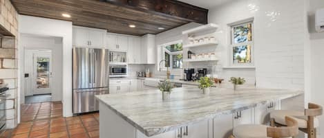 A modern kitchen with white cabinetry, a large island with bar stools, a stainless steel refrigerator, and a wooden ceiling.