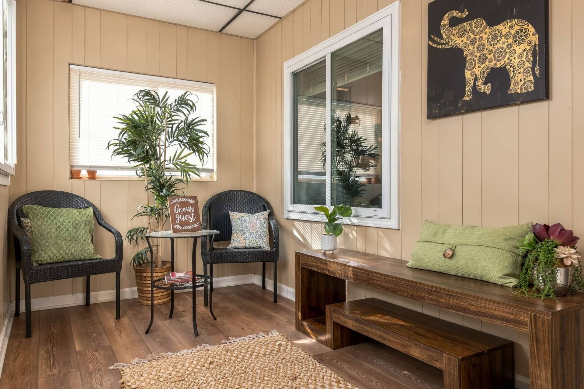 A cozy room with a wooden bench, two wicker chairs, a small table with a sign, and a large window with blinds.