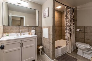 Ein Badezimmer mit weißem Waschbecken, Spiegel und Toilette, eine Dusche mit gemustertem Vorhang und ein kleiner Hocker.