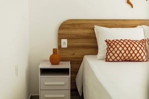 Una cama bien hecha con cabecera de madera, una mesita de noche blanca con cajones y un solo jarrón naranja sobre la mesita.