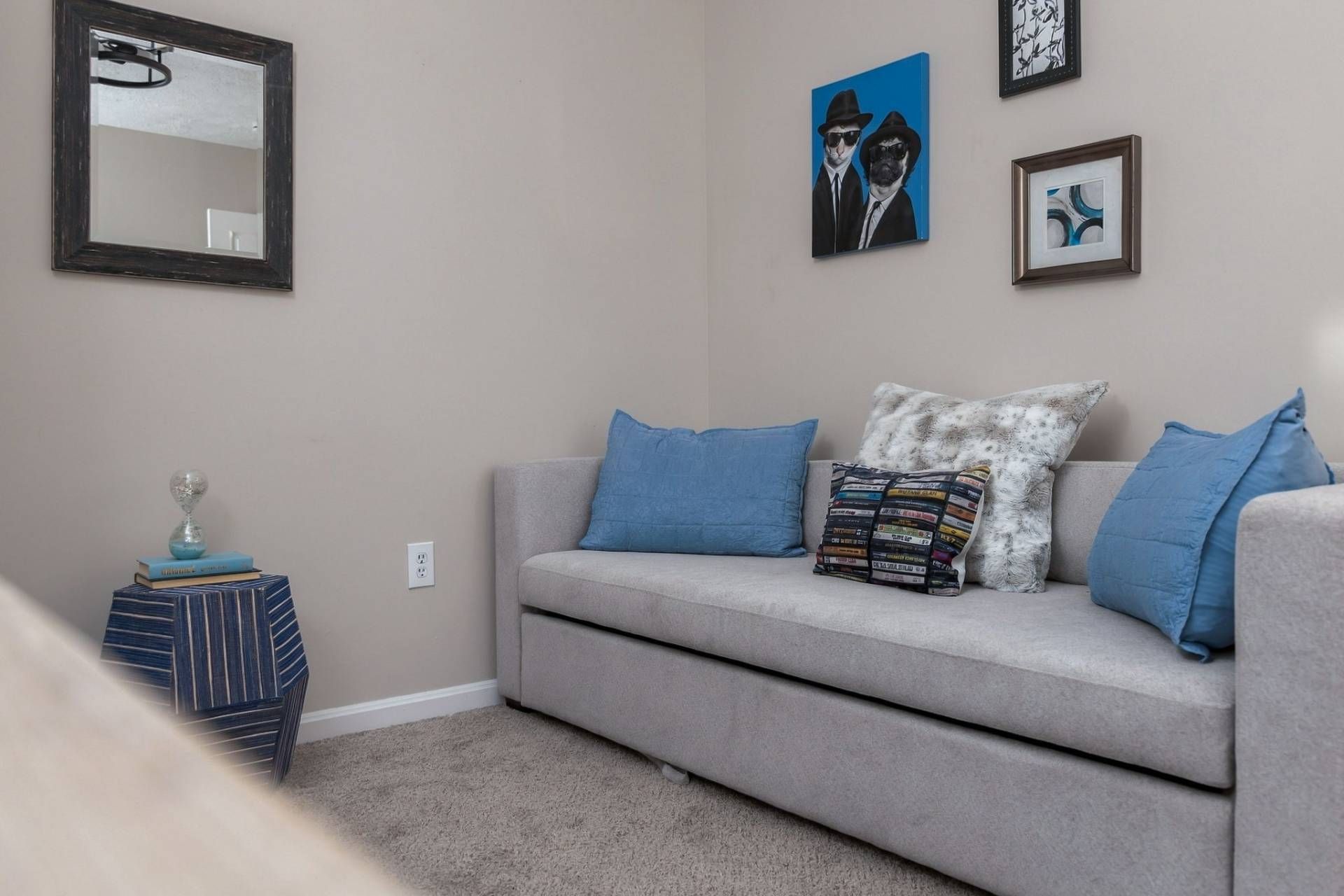 A living room with a grey sofa, blue cushions, a small side table with books, and a mirror on the wall.