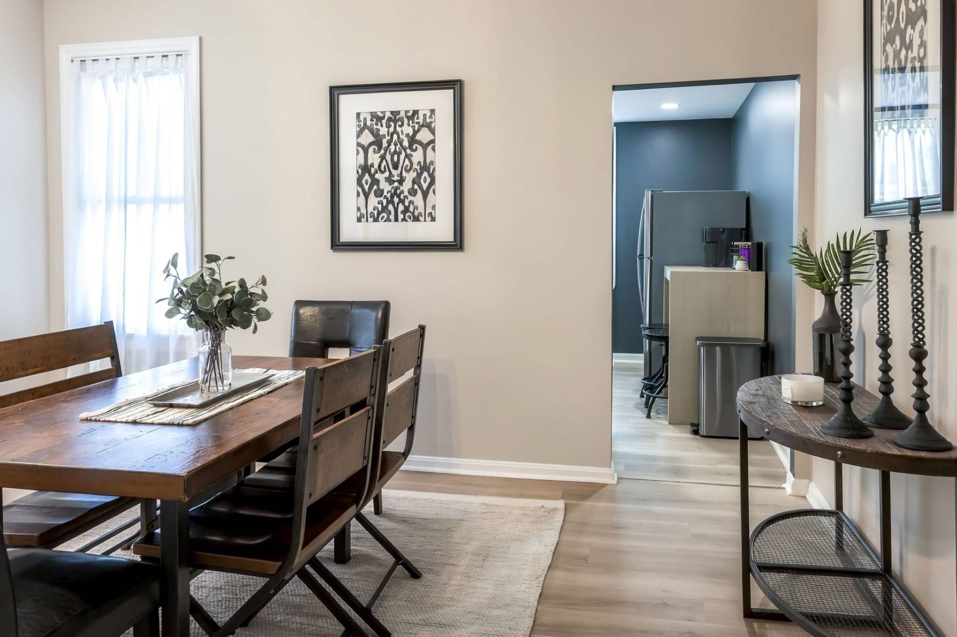 A dining area with a wooden table, chairs, and a vase of greenery. There is a framed artwork on the wall and a kitchen area in the background.