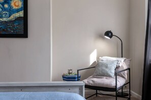 A bedroom with a bed, a chair with cushions, a bedside table with books, a lamp, and a painting on the wall. - Summit Row | Porch House (Pittsburgh)