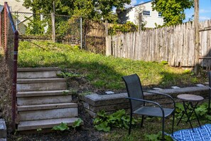 A backyard with a set of concrete steps leading up to a wooden fence, a metal chair, and a small round table. - Summit Row | Porch House (Pittsburgh)