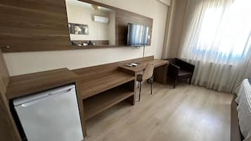 A hotel room with a wooden desk, a television, a small refrigerator, and a large window with curtains.