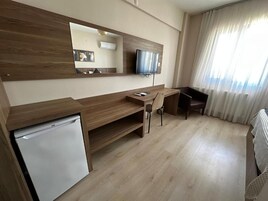 Habitación de hotel con escritorio de madera, televisor, refrigerador pequeño y ventana grande con cortinas.