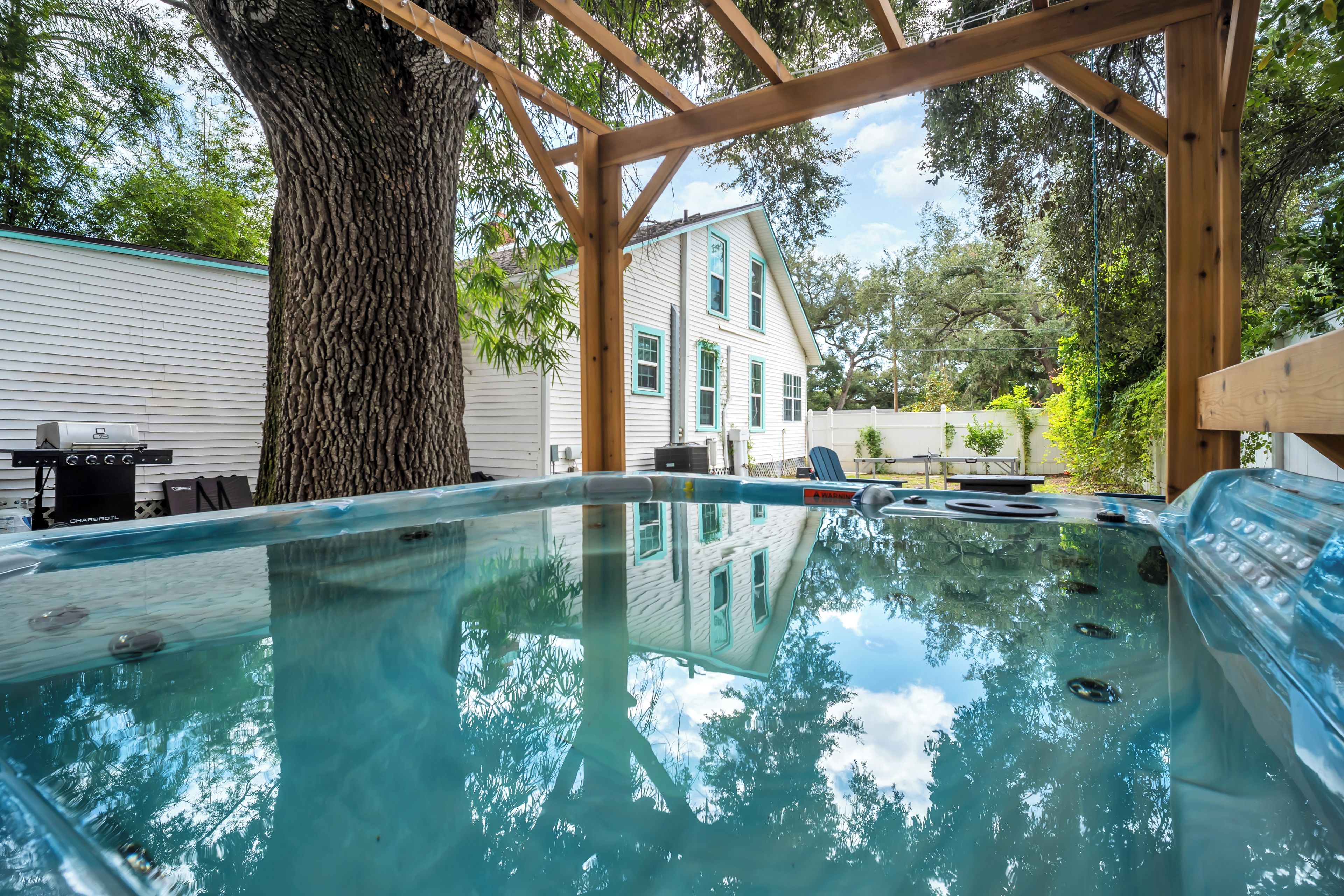 A hot tub under a wooden pergola with a tree and a house in the background.