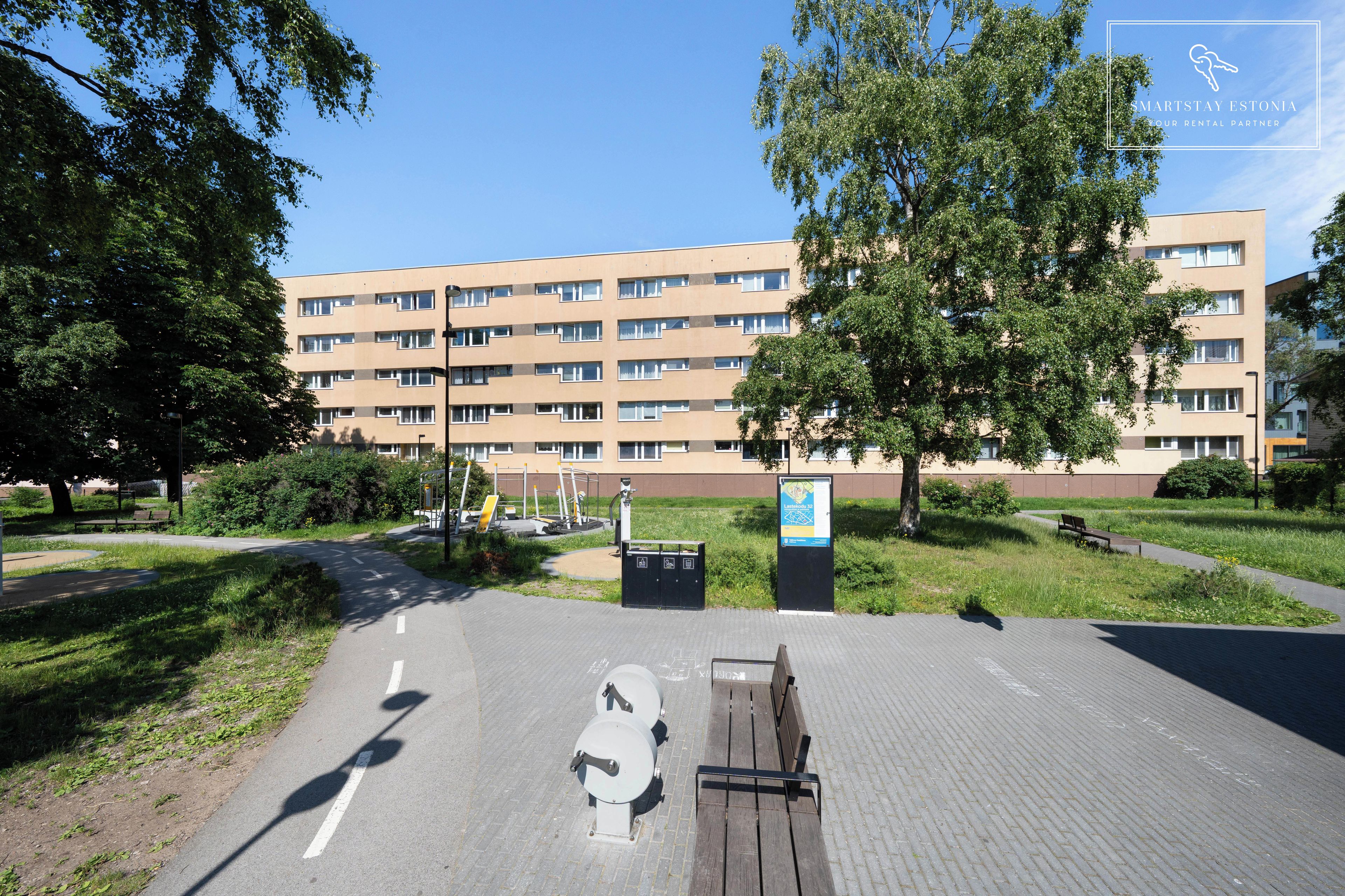 Un edificio de varios pisos con fachada plana, un área pavimentada con una banca y un cartel.