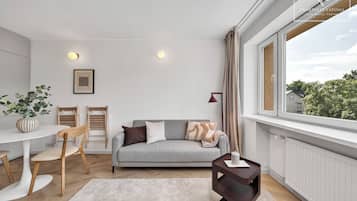 Un salón moderno con un sofá gris, sillas de madera, una mesita y vistas al exterior con vegetación.