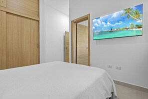 A bedroom with a bed, a wooden wardrobe, a door, and a wall-mounted TV displaying a beach scene. - 2BR Townhome w/Fast Wi-Fi, Backyard, Gazebo & Pool (Punta Cana)