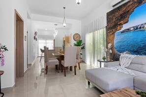 A modern living room with a sofa, dining table, chairs, and a large painting of a coastal scene. - 2BR Townhome w/Fast Wi-Fi, Backyard, Gazebo & Pool (Punta Cana)