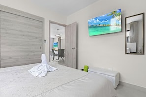 A hotel room with a large bed, a painting of a beach, a desk with a chair, and a bathroom mirror. - 3BR Villa w/ Patio, Pool Near Downtown & Beaches (Punta Cana)