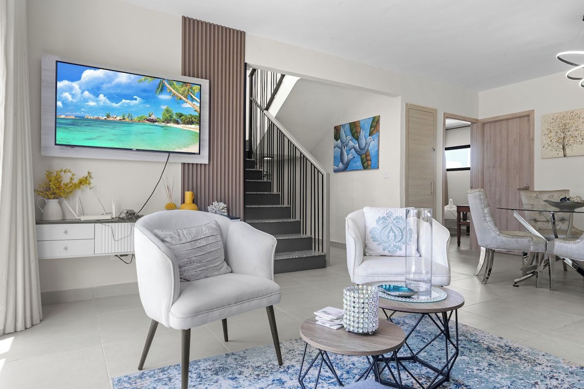 A modern living room with a flat-screen TV, a staircase, and a dining area.