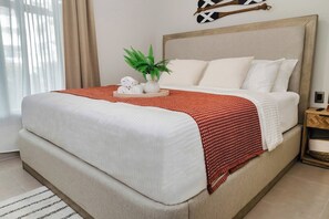 A neatly made bed with white linens and a red patterned throw, a small potted plant, and a tray with rolled towels on a wooden nightstand. - High-end 2BR w/Private Pool + BBQ + Steps to Beach (Punta Cana)