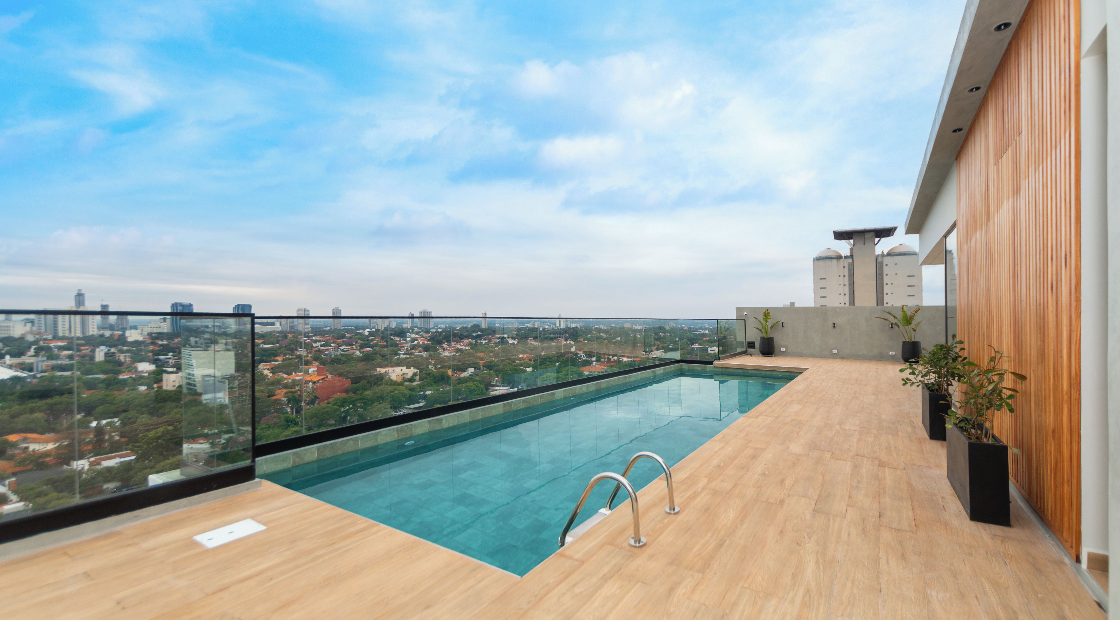 A rooftop pool with a glass barrier, wooden deck, and potted plants.
