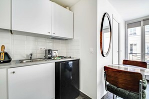 Eine kleine Küche mit weißen Schränken, einem schwarzen Kühlschrank, einer Kaffeemaschine und einem Toaster. Es gibt einen Esstisch mit einem Stuhl und einen großen Spiegel an der Wand.