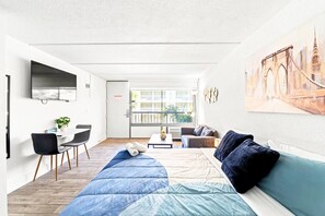 A modern hotel room with a bed, a seating area with a table and chairs, a TV mounted on the wall, and a large window offering a view of the city. - Near Disney + Pool + Gym + Park | Orl (Kissimmee)