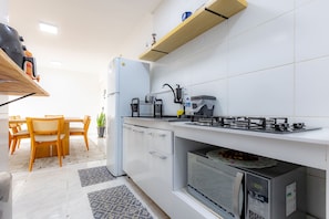 A compact kitchen with a refrigerator, stove, microwave, and a small dining area with a table and chairs. - Apto c Churrasqueira e Garagem - Jardim Iate Clube (Balneário Camboriú)
