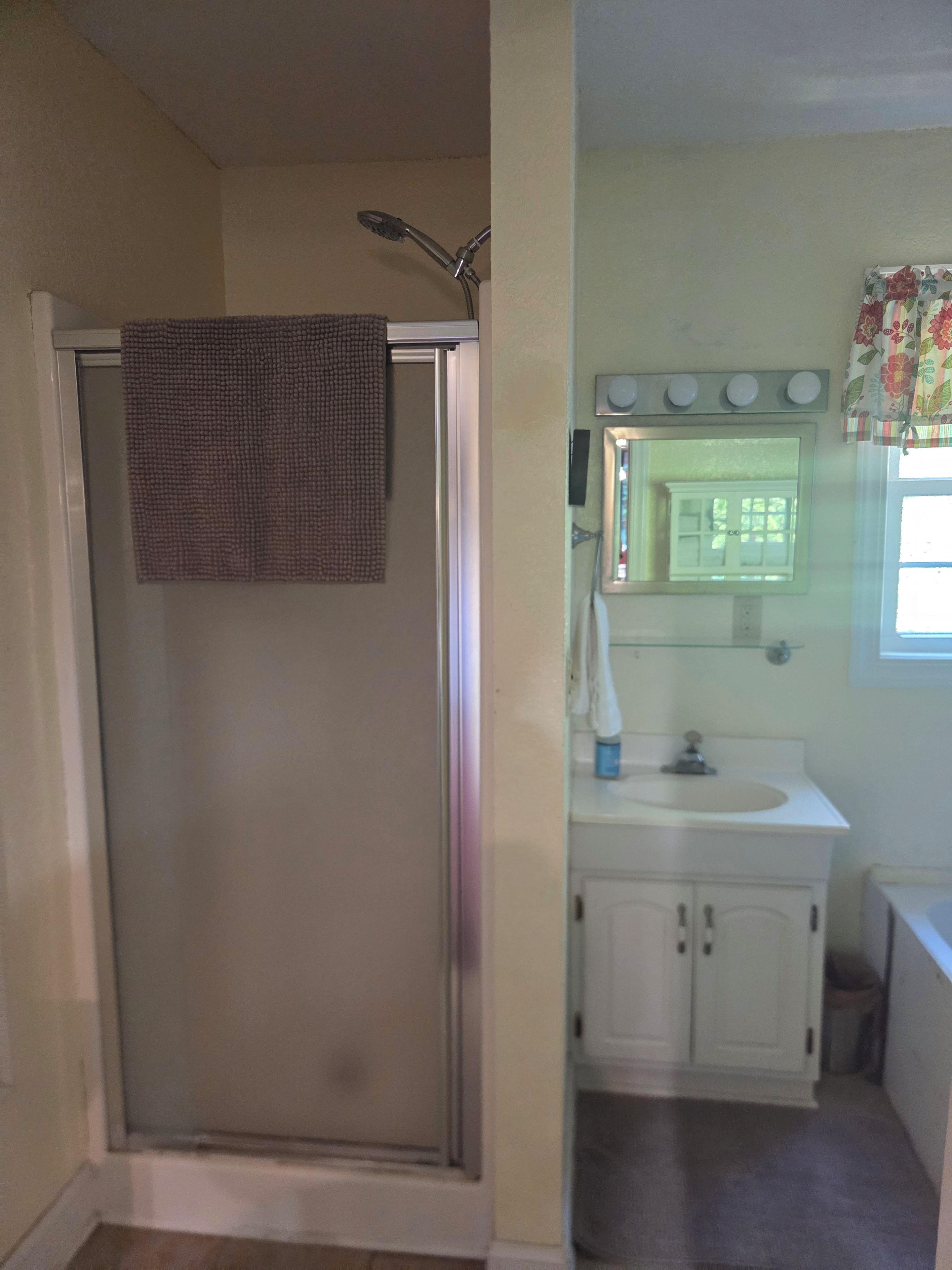 A bathroom with a glass-enclosed shower, a sink with a mirror above it, and a window with a floral curtain.