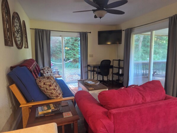 A living room with a red sofa, a blue armchair, a wooden coffee table with books, a ceiling fan, and a sliding glass door leading to a deck.