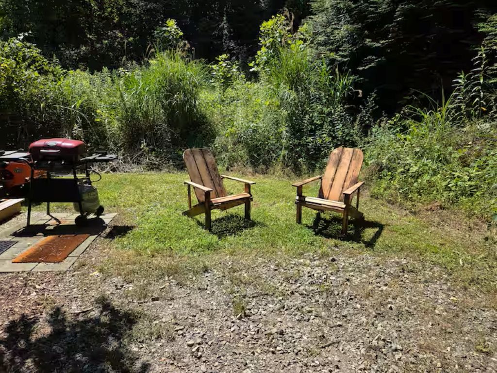 Dos sillas Adirondack de madera en un área con césped, una parrilla y un saco de carbón cerca.