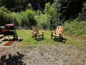 Two wooden Adirondack chairs in a grassy area with a grill and a bag of charcoal nearby.