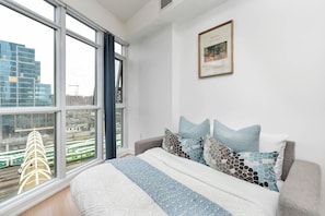 Ein Schlafzimmer mit großem Fenster, einem Bett mit gemusterten Betttextilien und einem gerahmten Bild an der Wand.