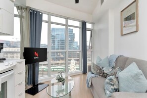 Ein modernes Wohnzimmer mit einem Flachbildfernseher, einer Couch mit gemusterten Kissen, einem Glastisch und großen Fenstern, die einen Blick auf die Stadt bieten.