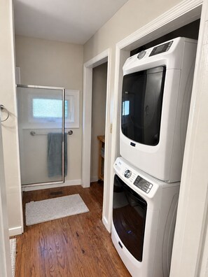 A laundry room with stacked washer and dryer units, a glass shower enclosure, and a wooden floor. - Restored Historic Home with Southern Porch Charm / Walk to Downtown Eufaula (Eufaula)