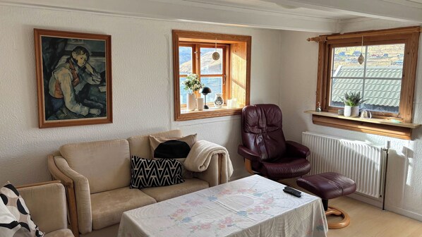 A cozy living room with a sofa, a leather armchair, a table with a floral tablecloth, and a painting on the wall.