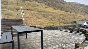 A wooden deck with a table and bench, overlooking a hilly landscape with a car parked nearby.