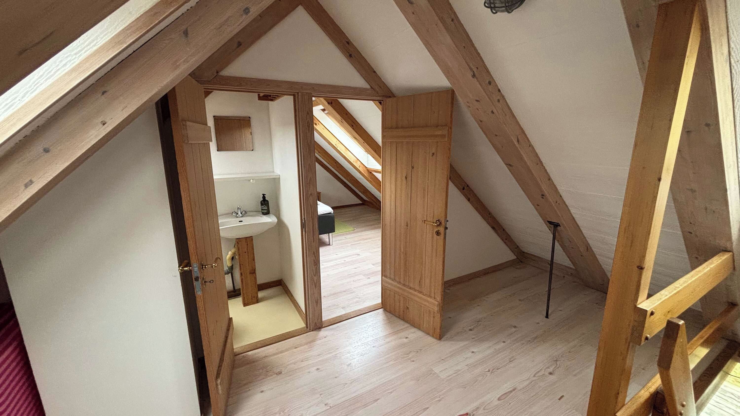 Una habitación con techo inclinado, vigas de madera, puerta de madera y un baño visible con lavamanos e inodoro.