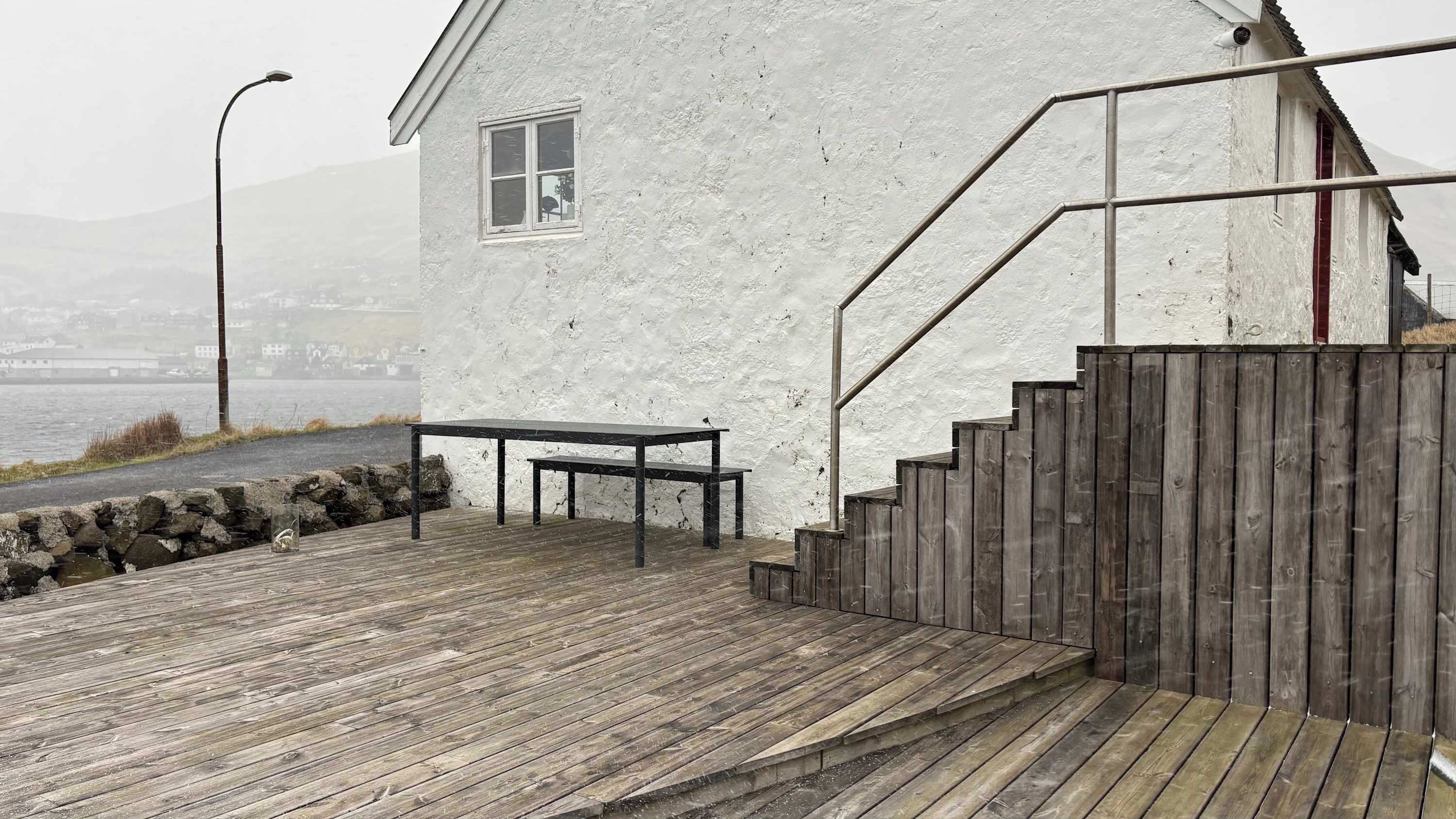 A wooden deck leading to a white building with a window and a bench.