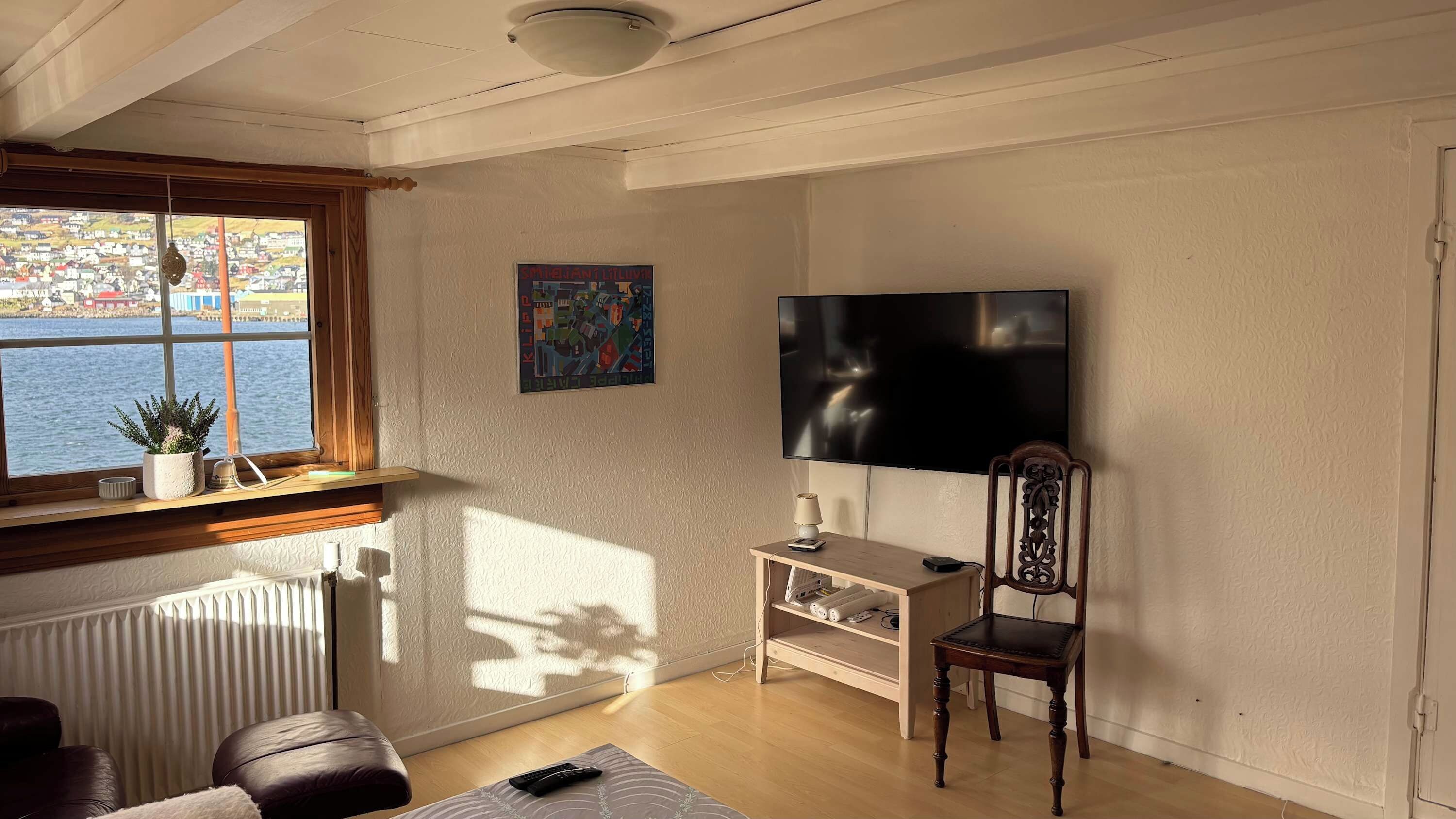 A living room with a large window, a flat-screen TV, a wooden chair, a small table, and a radiator.