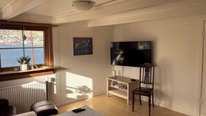 Ein Wohnzimmer mit großem Fenster, Flachbildfernseher, Holzstuhl, kleinem Tisch und Heizkörper.