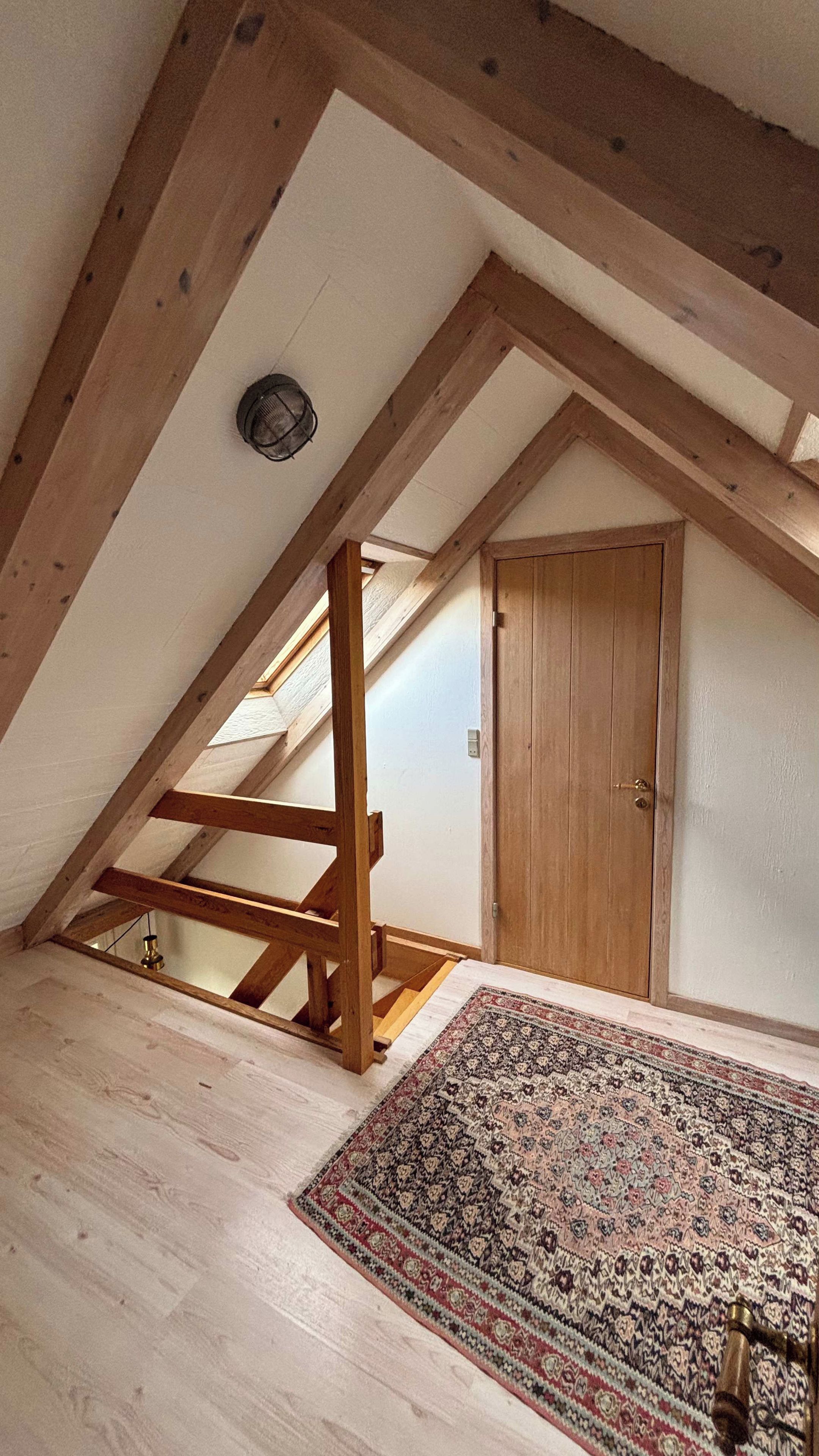 Ein Zimmer mit schrägverdeckter Decke, Holzbalken, einer Tür und einem gemusterten Teppich.