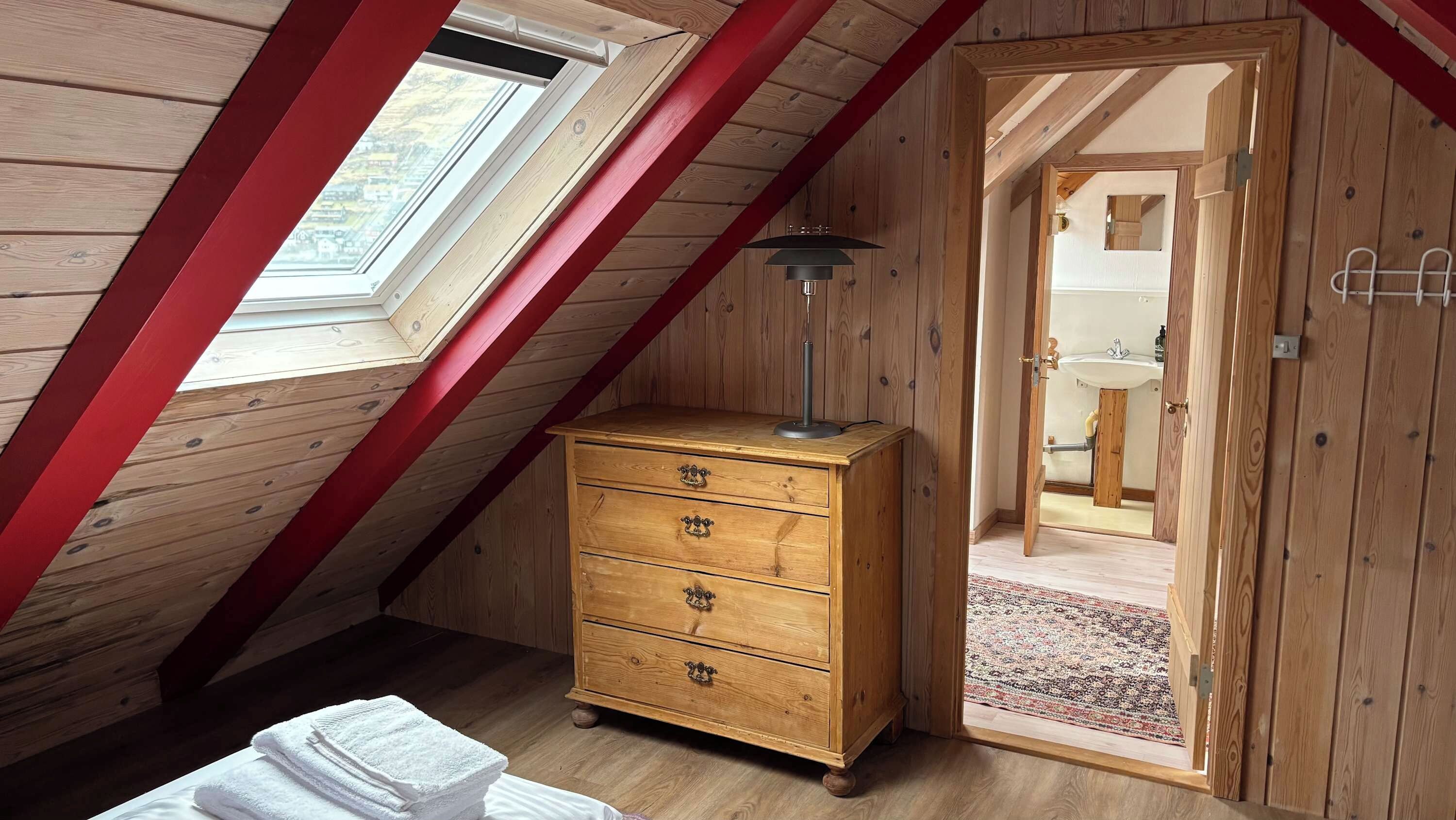 A room with a sloped ceiling, wooden walls, a wooden dresser with drawers, a small table with a lamp, and a view through a door to a bathroom.