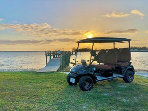 A golf cart parked on a grassy area near a dock with a sunset in the background. - Take in sweeping panoramic views from this 3-bed, 3-bath penthouse in the heart of Sebastian. (Sebastian)