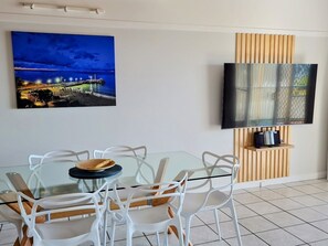 Een eethoek met een glazen tafel en witte stoelen, een flatscreen televisie bevestigd aan een houten paneel en een schilderij van een kustlandschap.